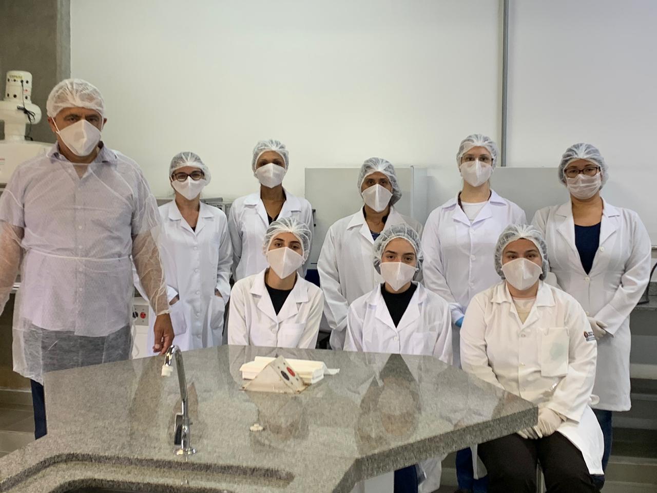 #PraCegoVer visualiza-se a imagem de nove profissionais e estudantes de farmácia na foto, todos vestidos com roupas de proteção e pousando para a foto no laboratório em que estão trabalhando.